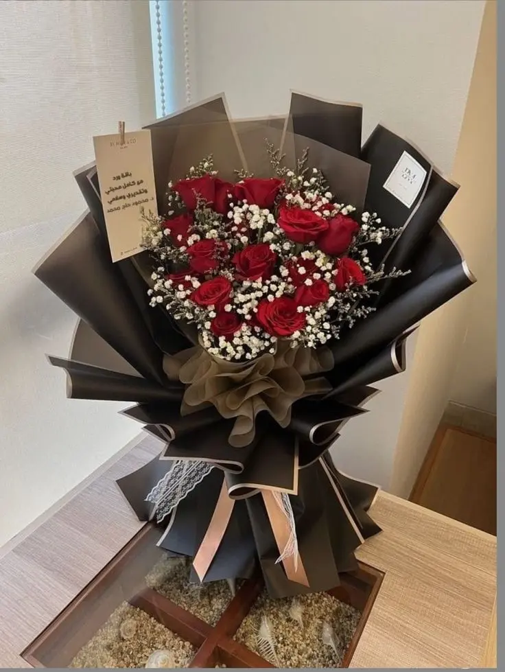 A bouquet of vibrant red roses accented with white baby's breath wrapped in elegant brown paper, tied with ribbon, and placed on a wooden table.