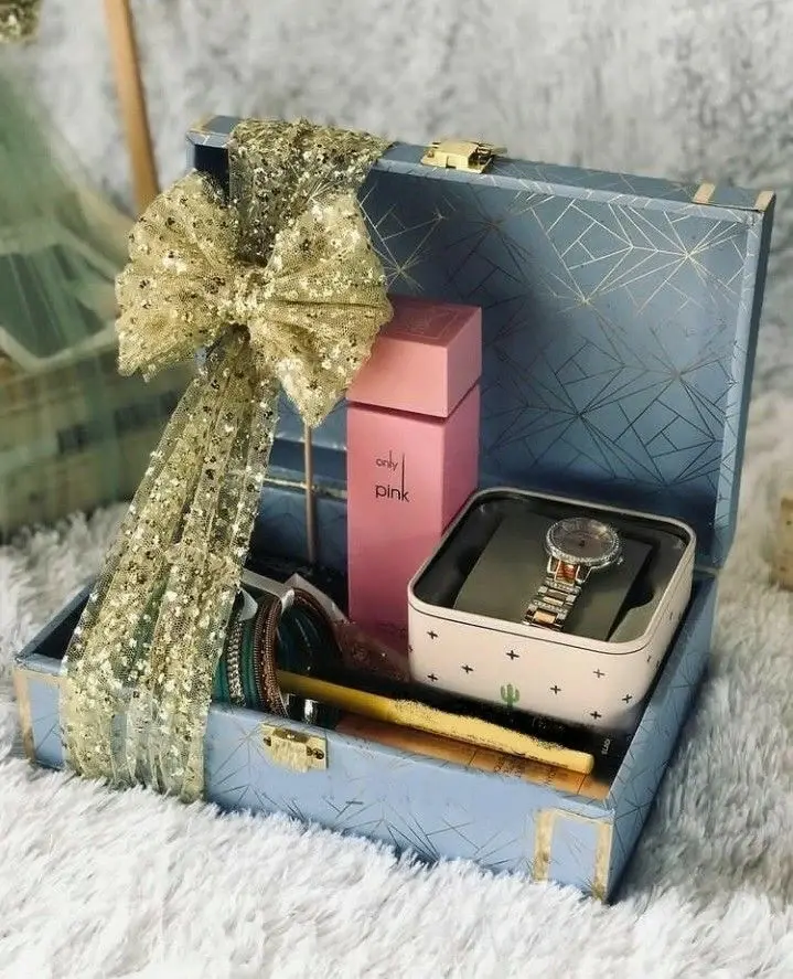 A blue gift box with a gold ribbon holds a watch, pink perfume box, bangles, and a rolled scroll. The setting is cozy, suggesting a festive or celebratory mood.